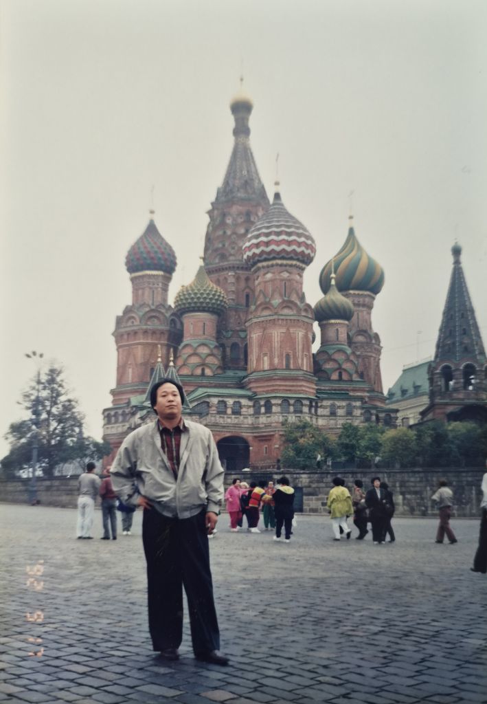 Faded color photograph of a man in a grey jacket standing in front of Saint Basil's Cathedral in Moscow on an overcast day.